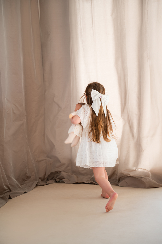 Kind in heller Kleidung tanzt barfuß mit Kuscheltier vor Stoffkulisse – natürliche People- und Lifestylefotografie mit sanftem Licht