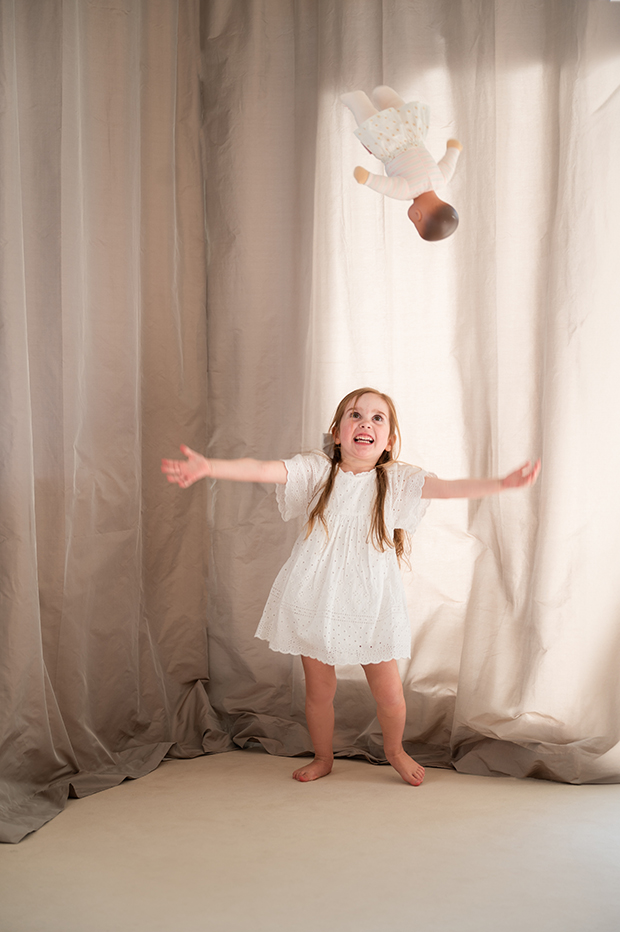 Kind in heller Kleidung tanzt barfuß mit Kuscheltier vor Stoffkulisse – natürliche People- und Lifestylefotografie mit sanftem Licht