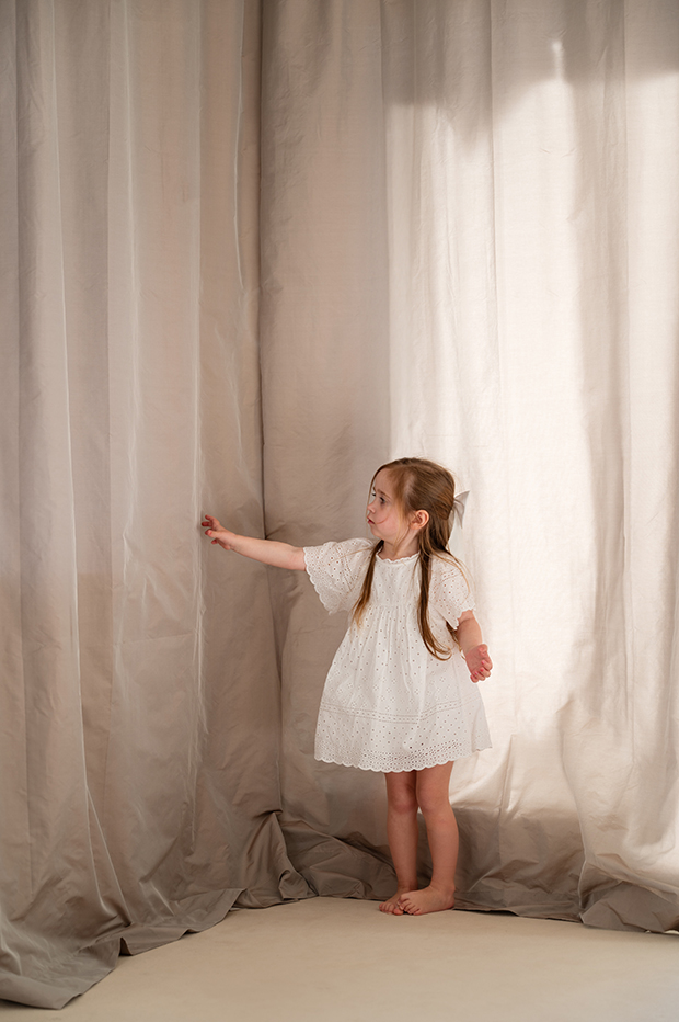 Kind in heller Kleidung tanzt barfuß mit Kuscheltier vor Stoffkulisse – natürliche People- und Lifestylefotografie mit sanftem Licht